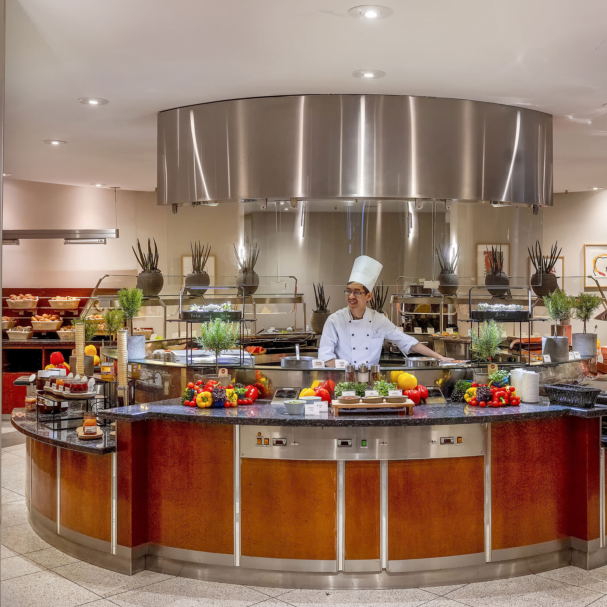 Front cooking Küchenchef an der Show-Cooking-Station im Maritim proArte Hotel Berlin, umgeben von frischem Gemüse und Kräutern.