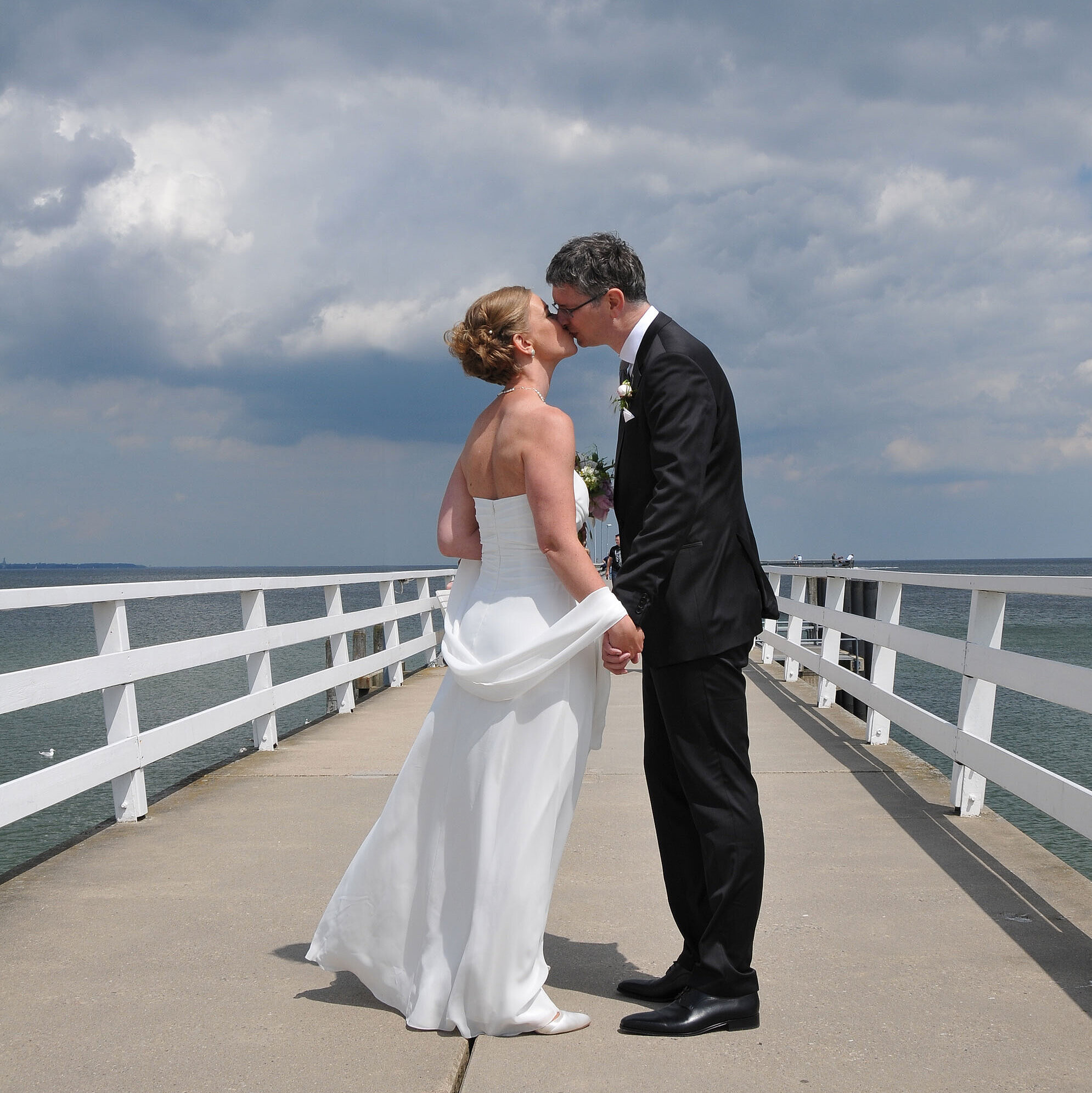 Hochzeitspaar auf der Seebrücke vor dem Maritim Hotel Timmendorfer Strand, das sich bei bewölktem Himmel einen Kuss gibt.