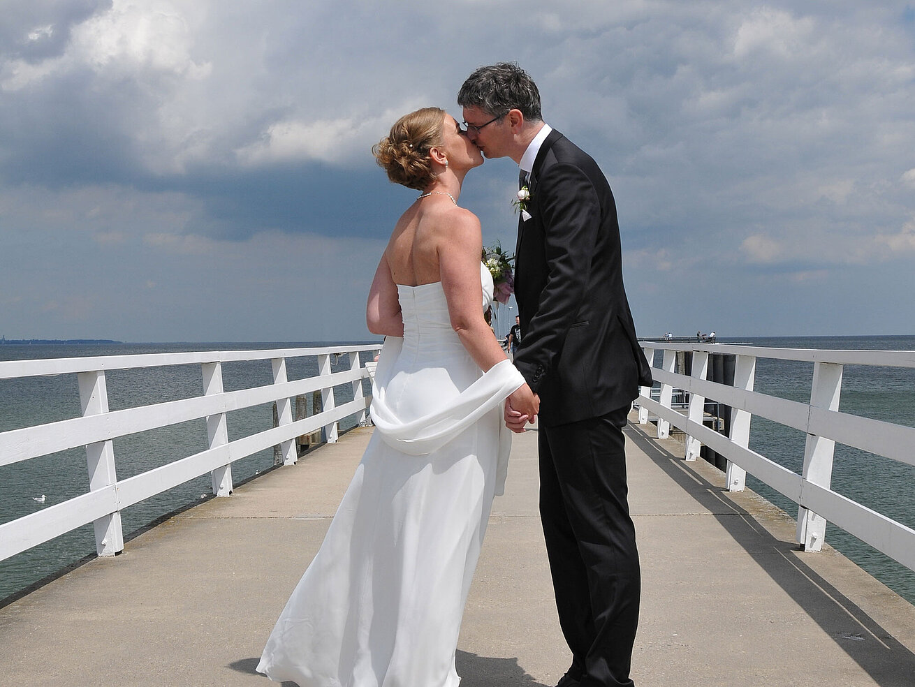 Hochzeit am Meer Hochzeitspaar auf der Seebrücke vor dem Maritim Hotel Timmendorfer Strand, das sich bei bewölktem Himmel einen Kuss gibt.