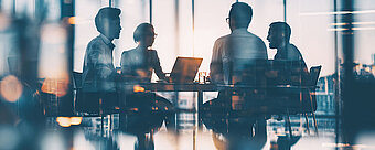 Vier Personen sitzen in einem modernen Konferenzraum mit großen Fenstern und besprechen sich am Tisch.