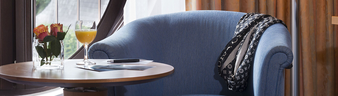  Gemütlicher Sitzbereich im Classic Zimmer des Maritim Hotel Fulda mit blauem Sessel, rundem Fenster, Blumen und Orangensaft.