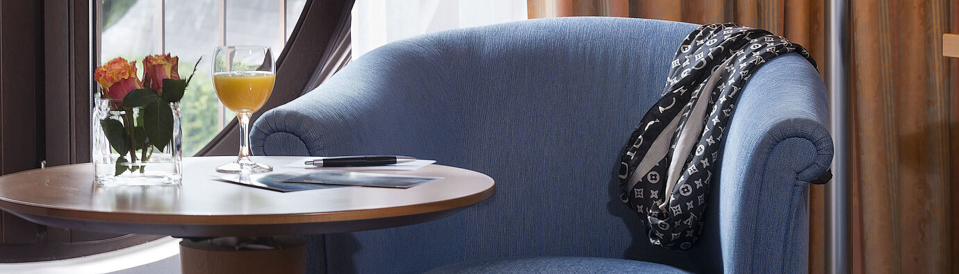 Zimmerdetail Gemütlicher Sitzbereich im Classic Zimmer des Maritim Hotel Fulda mit blauem Sessel, rundem Fenster, Blumen und Orangensaft.