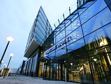 "Moderne Außenansicht des Maritim Hotel Düsseldorf mit gläserner Fassade und leuchtendem Hotellogo bei Dämmerung.