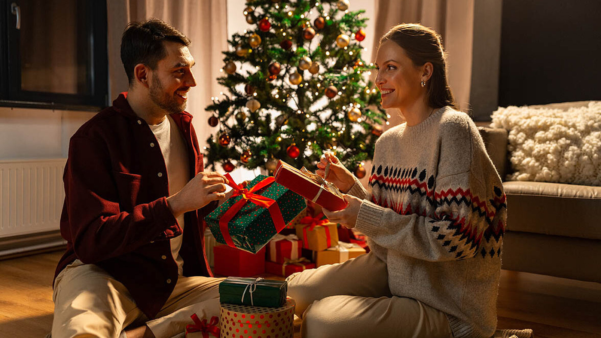 Zwei Personen sitzen vor einem geschmückten Weihnachtsbaum und tauschen liebevoll verpackte Geschenke aus.