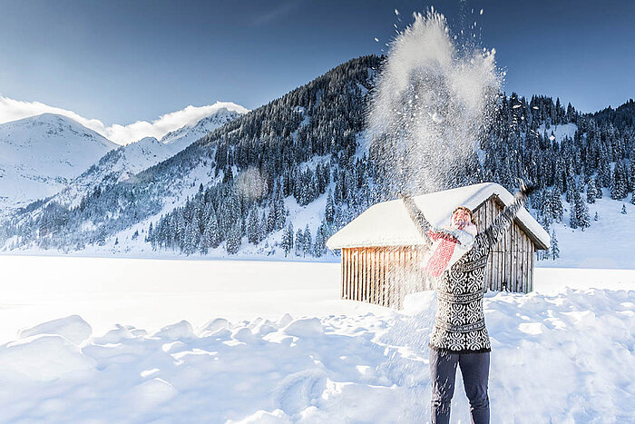 © mmphoto - AdobeStock.com Person wirft Schnee in die Luft vor verschneiten Bergen und einer Hütte in strahlender Winterlandschaft.
