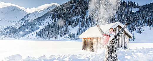 © mmphoto - AdobeStock.com Person wirft Schnee in die Luft vor verschneiten Bergen und einer Hütte in strahlender Winterlandschaft.