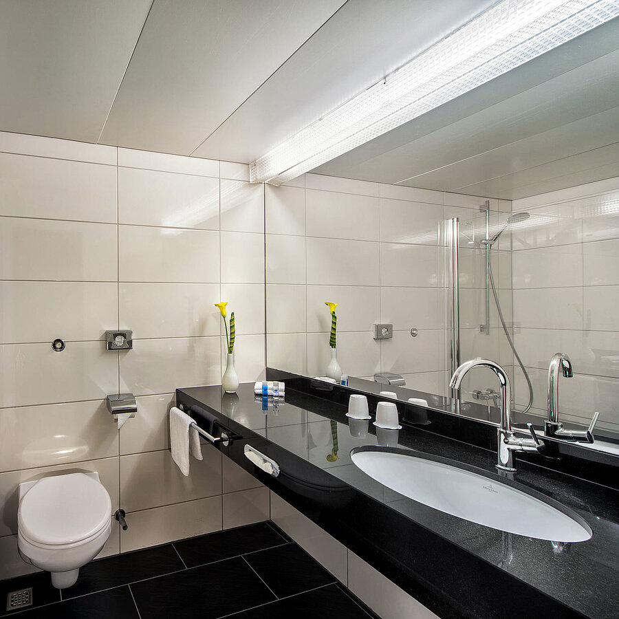 Badezimmer im Maritim Hotel Kiel mit Badewanne, Glasduschwand, Waschbecken und großem Spiegel auf schwarzer Ablage.