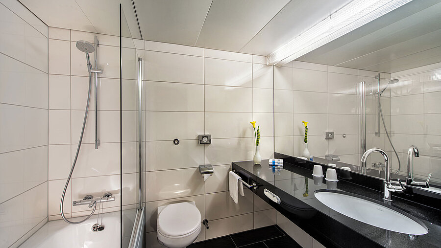 Badezimmer im Maritim Hotel Kiel mit Badewanne, Glasduschwand, Waschbecken und großem Spiegel auf schwarzer Ablage.