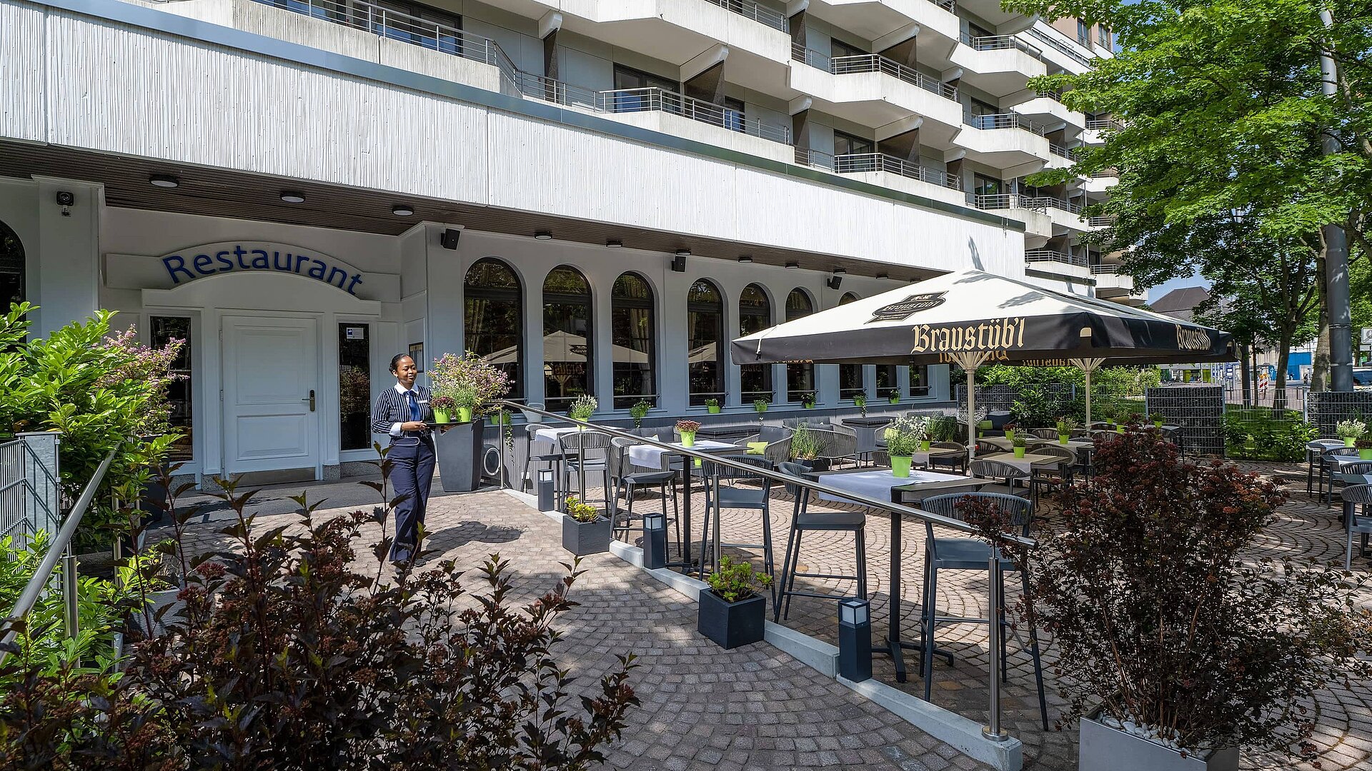 Sommerliche Restaurantterrasse des Maritim Hotel Darmstadt mit Außenfassade und Tischen unter einem Sonnenschirm