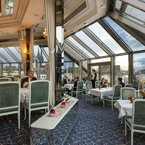 Elegantes Restaurant mit Glasdach und Blick auf den Kölner Dom und die Hohenzollernbrücke, Gäste genießen feines Essen.