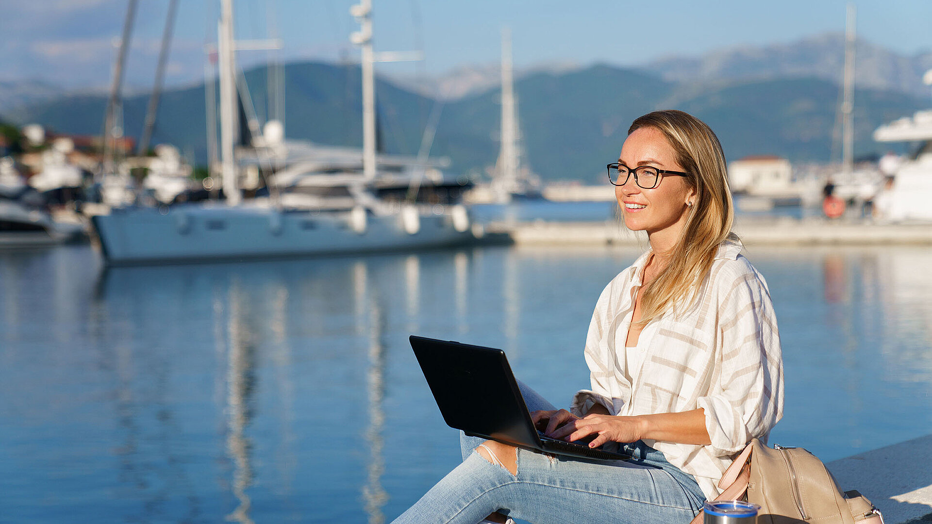 © Mary Anlens - AdobeStock.com Frau arbeitet mit Laptop am Hafen vor Segelbooten und ruhigem Wasser bei Sonnenschein.