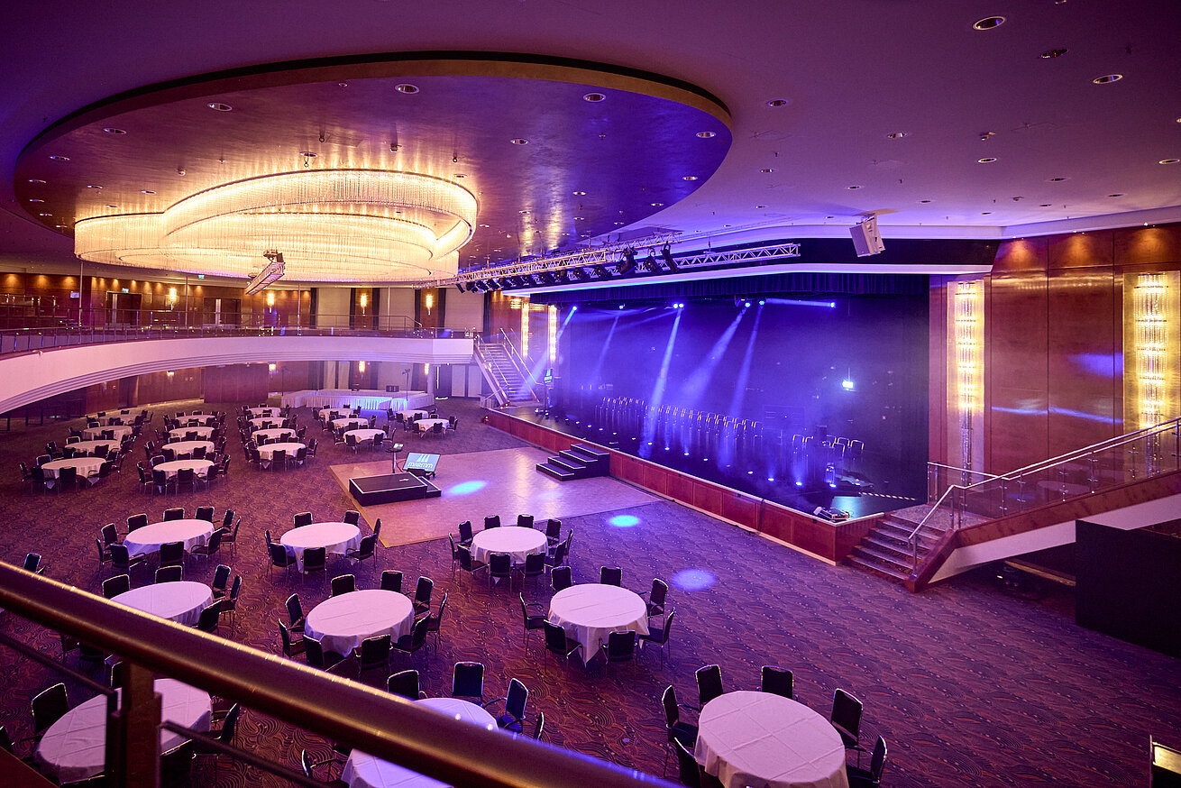 Großer Ballsaal mit Bühne, runden Tischen und moderner Lichttechnik für Gala oder Tagung.