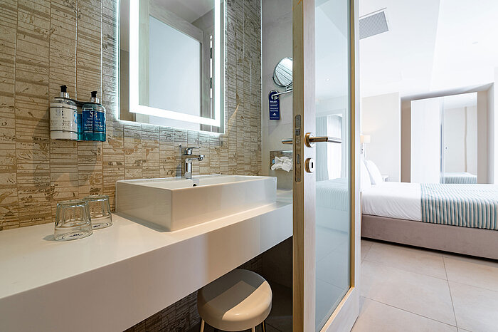 Modernes Badezimmer der Junior Suite im Maritim Antonine Hotel & Spa Malta mit beleuchtetem Spiegel, stilvollem Waschtisch und Blick in den hellen Schlafbereich.
