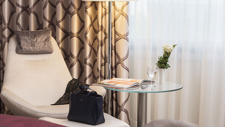 Detail eines Classic Zimmers im Maritim Hotel Bonn mit gemütlichem Sessel, Vorhang, Tisch, Glas, Rose und einer Tasche.