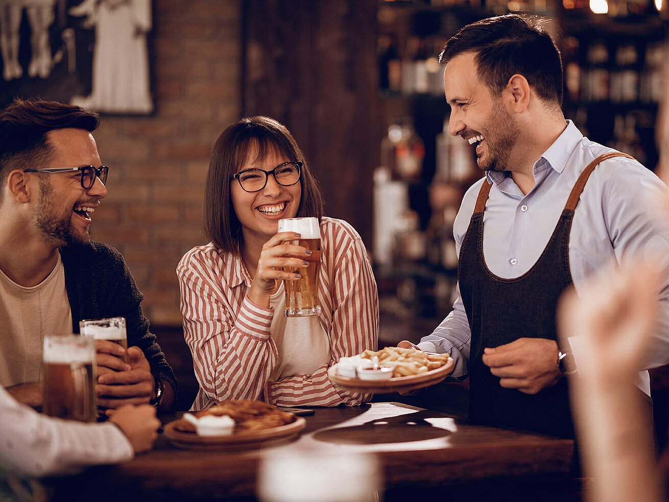 © Drazen - AdobeStock.com Lachende Gäste genießen Bier und Snacks, bedient von freundlichem Kellner in Bar