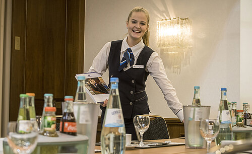 Freundliche Servicemitarbeiterin im Salon Lenné des Maritim Hotels Bonn mit Getränken und Tagungsunterlagen auf dem Tisch.