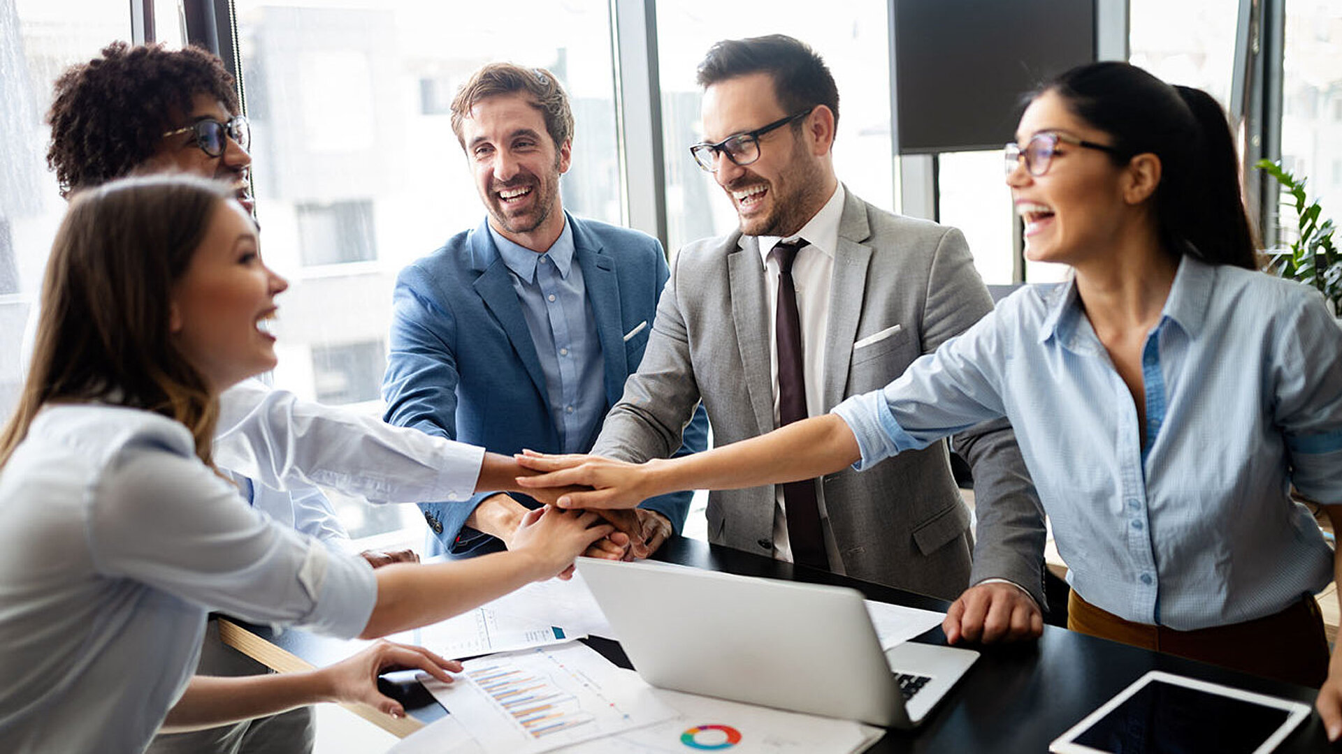 Motiviertes Business-Team im Büro legt Hände zusammen, Meeting mit Laptop und Unterlagen am Konferenztisch