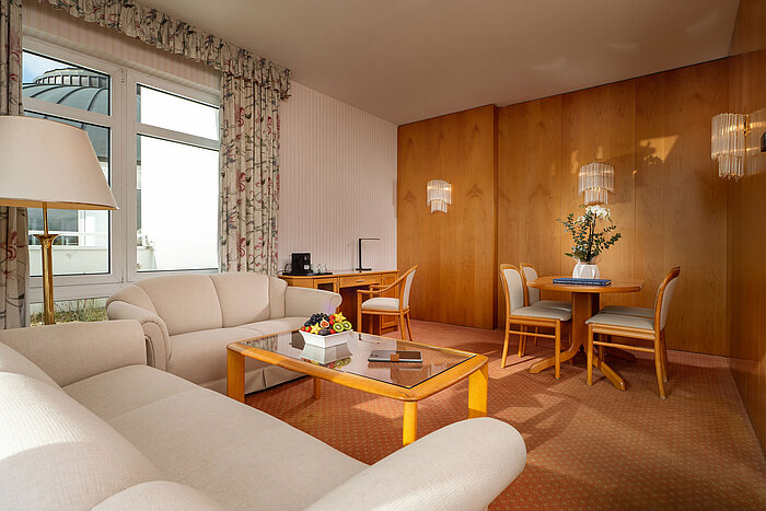 Junior Suite mit hellem Wohnbereich, Sofa, Glastisch, Essplatz und gemütlicher Einrichtung im Maritim Hotel.