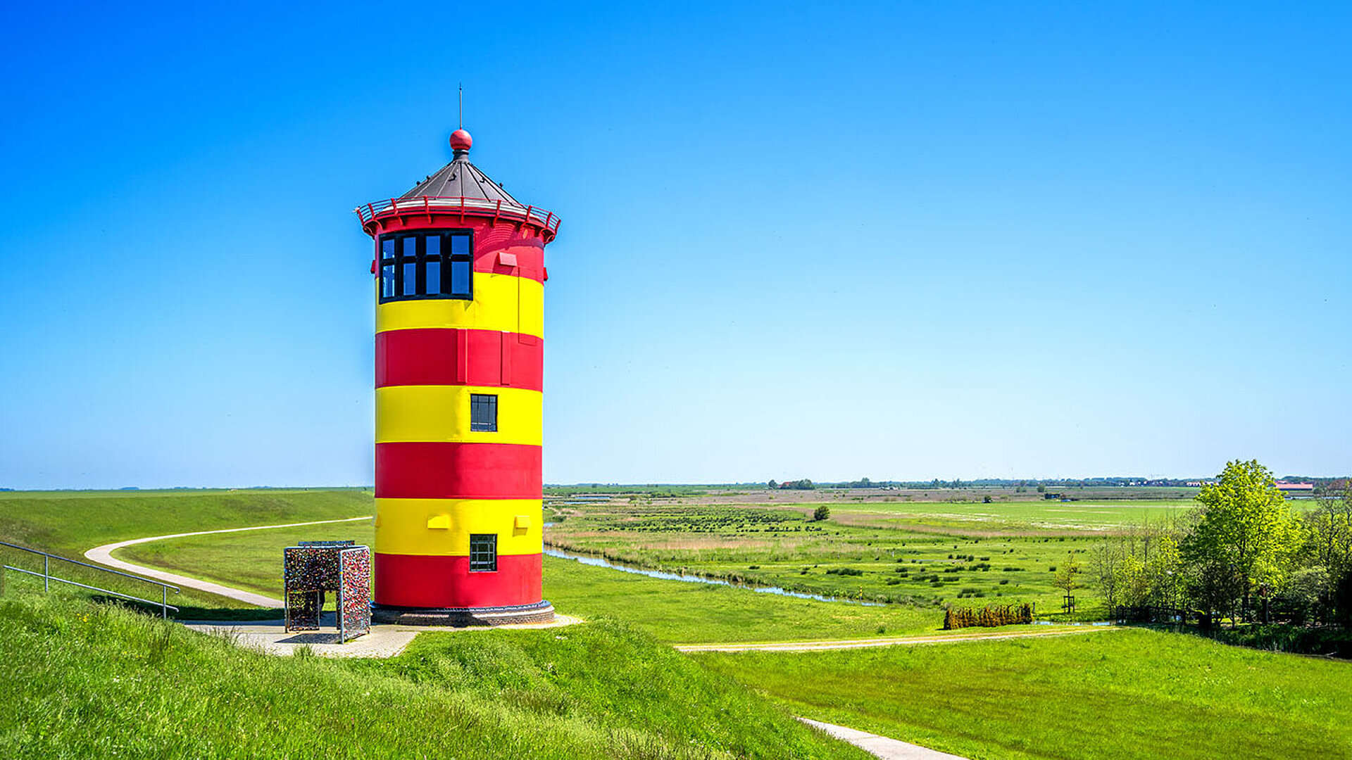 © Sina Ettmer - AdobeStock.com Gelb-rot gestreifter Leuchtturm umgeben von Grün in Pilsum in Ostfriesland