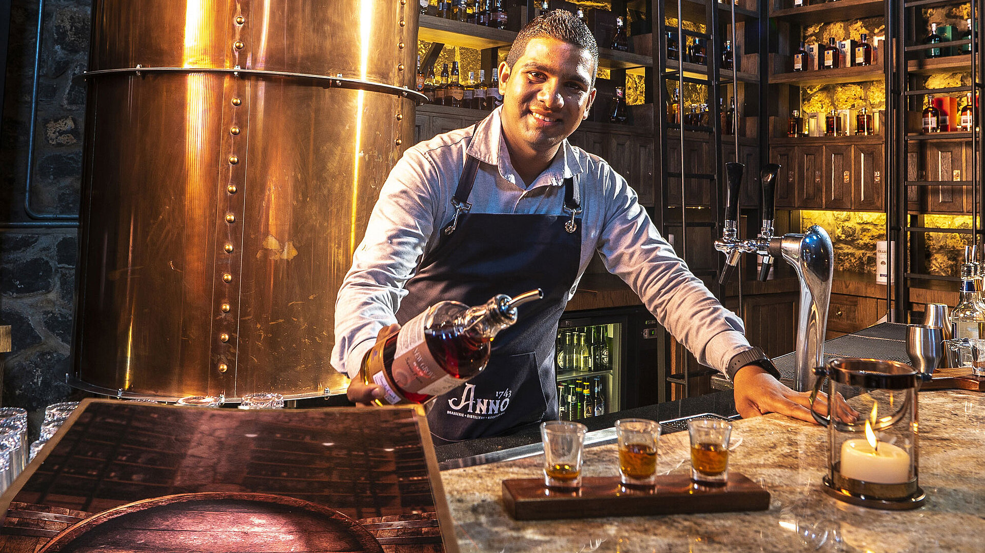 "The Distillery" Barkeeper serviert Spirituosen an der Bar mit Kupferdestille und beleuchteten Regalen