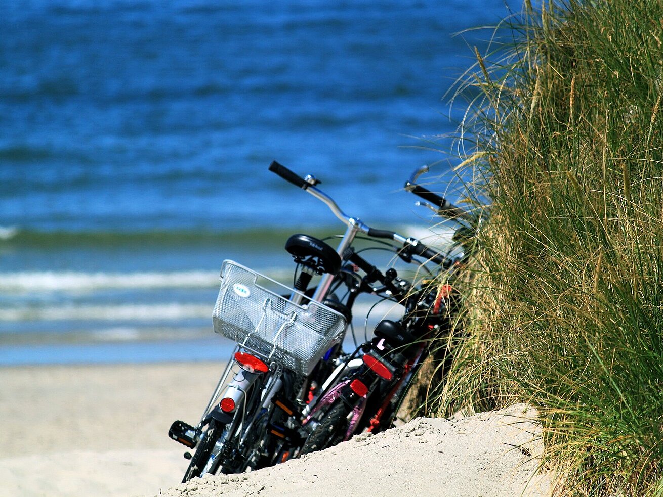 Zwei Fahrräder parken hinter einer Düne an einem sandigen Strand mit blauem Meer im Hintergrund.