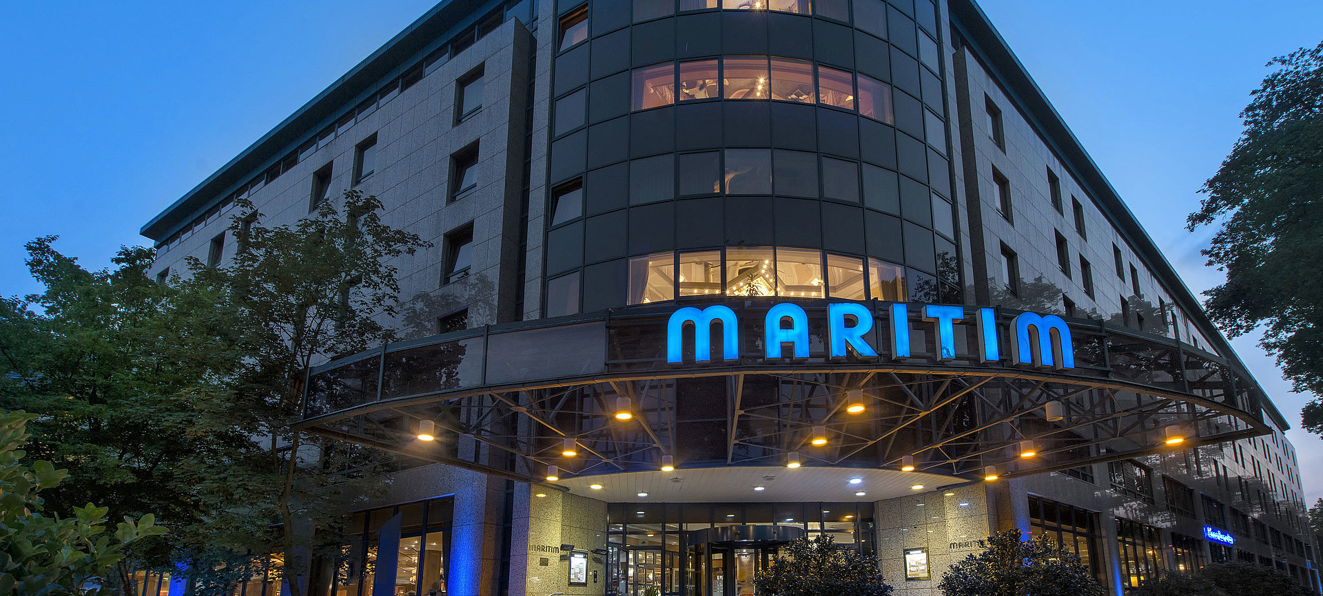 Außenansicht des Maritim Hotel Bremen bei Dämmerung, beleuchtetes Logo und moderne Architektur.