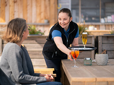 Eine Kellnerin serviert Aperol Spritz und Bier an einen Gast auf der Terrasse der Beach Lounge, mit Strand im Hintergrund.
