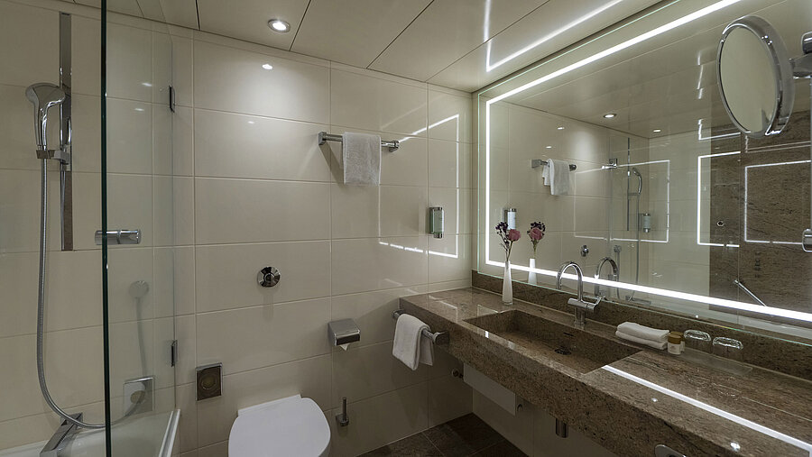 Modernes Badezimmer im Maritim Hotel Bremen mit beleuchtetem Spiegel, Badewanne und hochwertigen Granitoberflächen.
