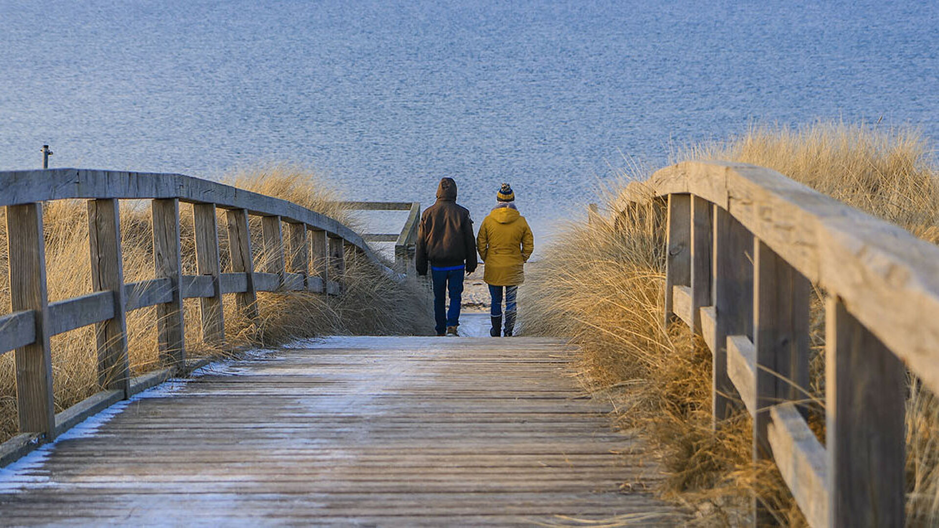 © Lars Gieger - AdobeStock.com Paar beim Winterspaziergang am Strand über Holzsteg Richtung Meer
