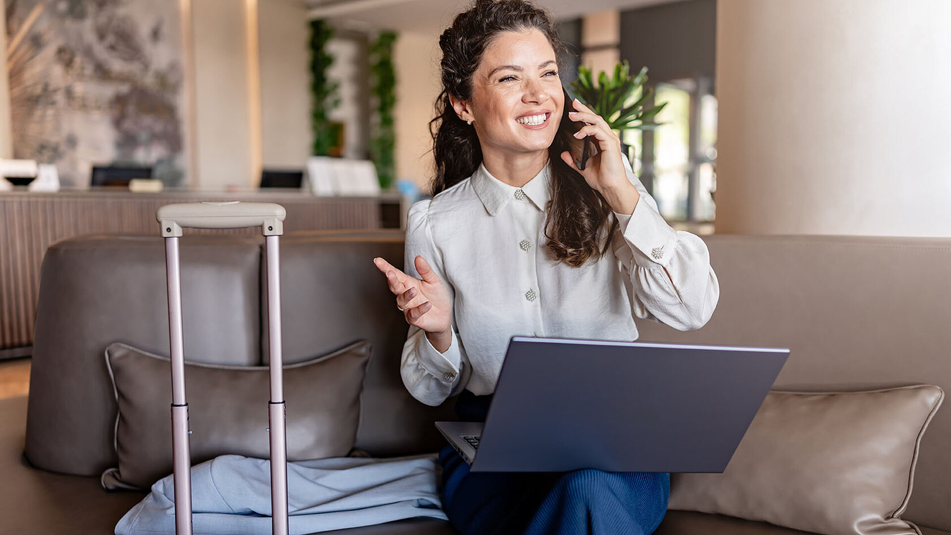 © lordn - AdobeStock.com Geschäftsfrau sitzt mit Laptop auf dem Sofa in einer Hotel-Lobby, telefoniert lächelnd und hat einen Koffer neben sich