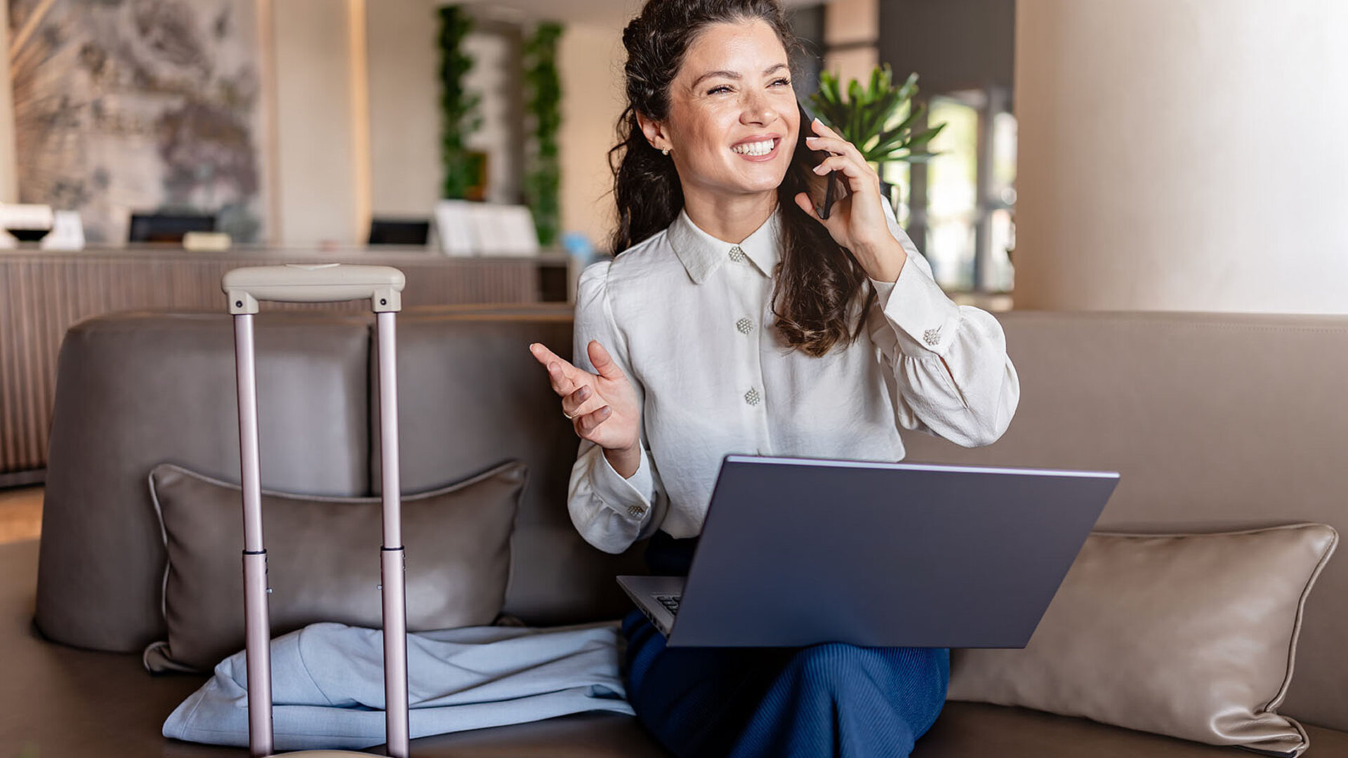 © lordn - AdobeStock.com Geschäftsfrau sitzt mit Laptop auf dem Sofa in einer Hotel-Lobby, telefoniert lächelnd und hat einen Koffer neben sich