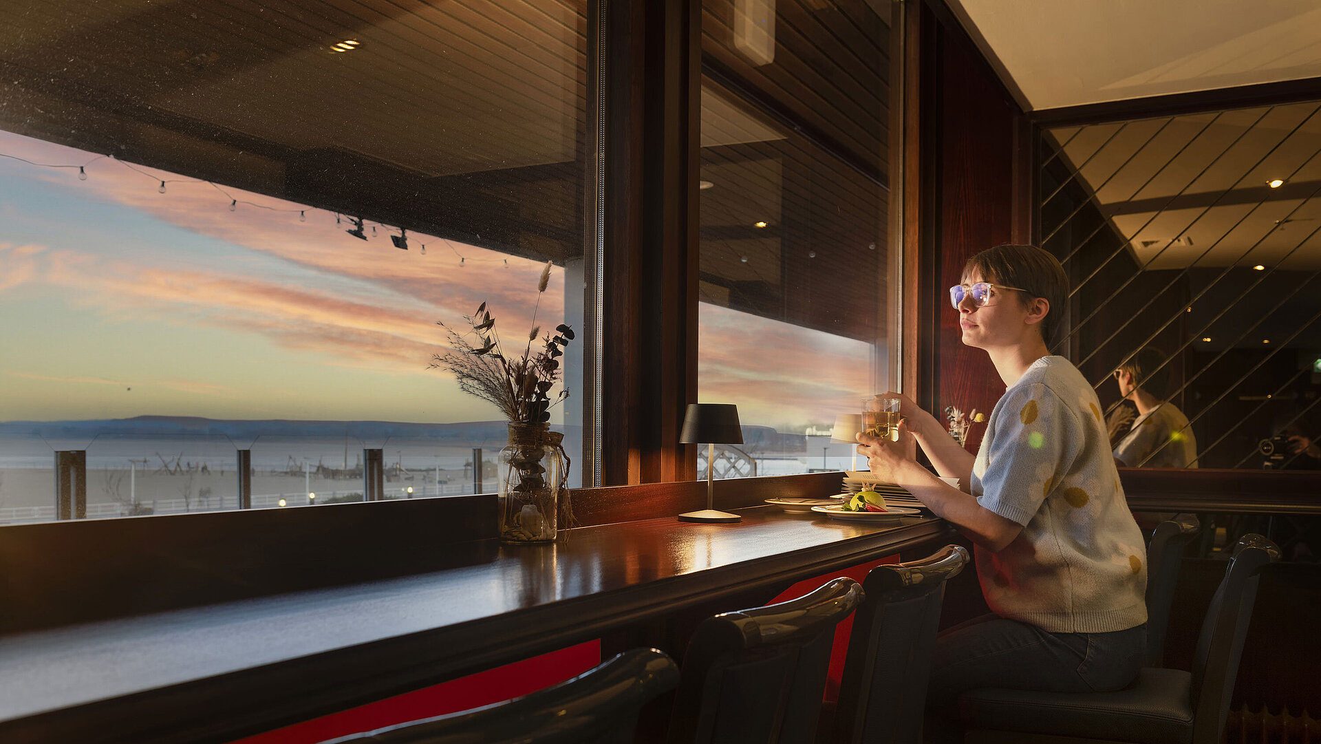 Gast sitzt mit Getränk am Fenster und blickt auf den Sonnenuntergang im Maritim Hotel Travemünde