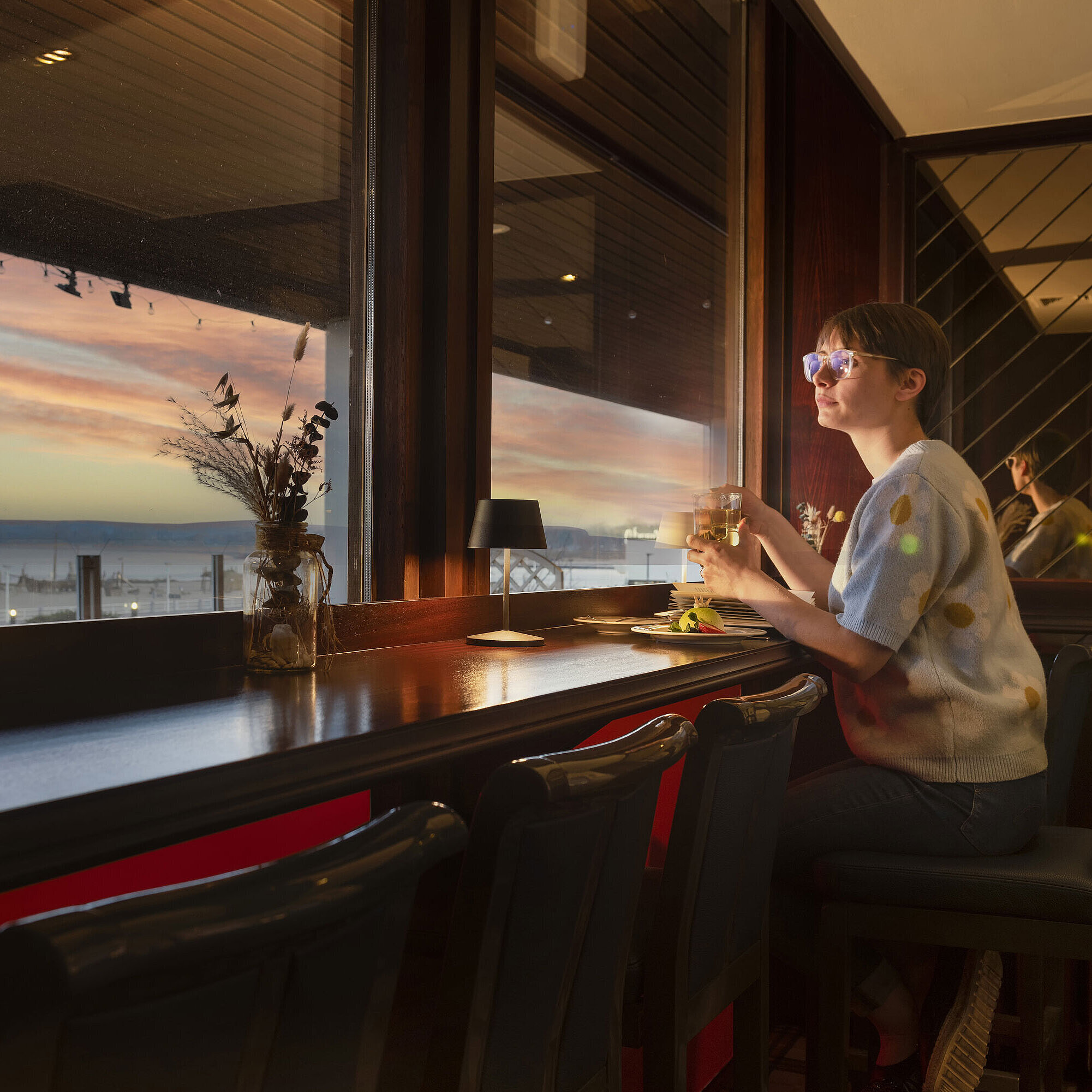 Gast sitzt mit Getränk am Fenster und blickt auf den Sonnenuntergang im Maritim Hotel Travemünde