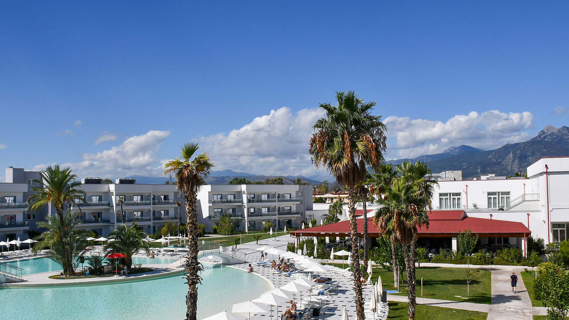 Panorama des Maritim Resort Calabria mit Pool, Palmen, weißen Gebäuden und Bergkulisse im Hintergrund bei klarem Himmel.