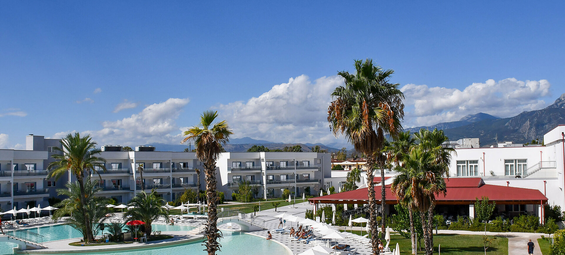 Panorama des Maritim Resort Calabria mit Pool, Palmen, weißen Gebäuden und Bergkulisse im Hintergrund bei klarem Himmel.