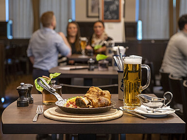 Servierter Schweinebraten mit Bier im Restaurant des Maritim Hotel Düsseldorf, im Hintergrund Gäste in geselliger Atmosphäre.