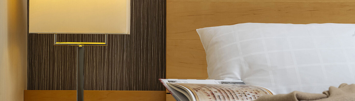 Detailaufnahme eines Nachttisches im Maritim Hotel Düsseldorf mit Lampe, Glas Wasser und Buch auf einem Bett.