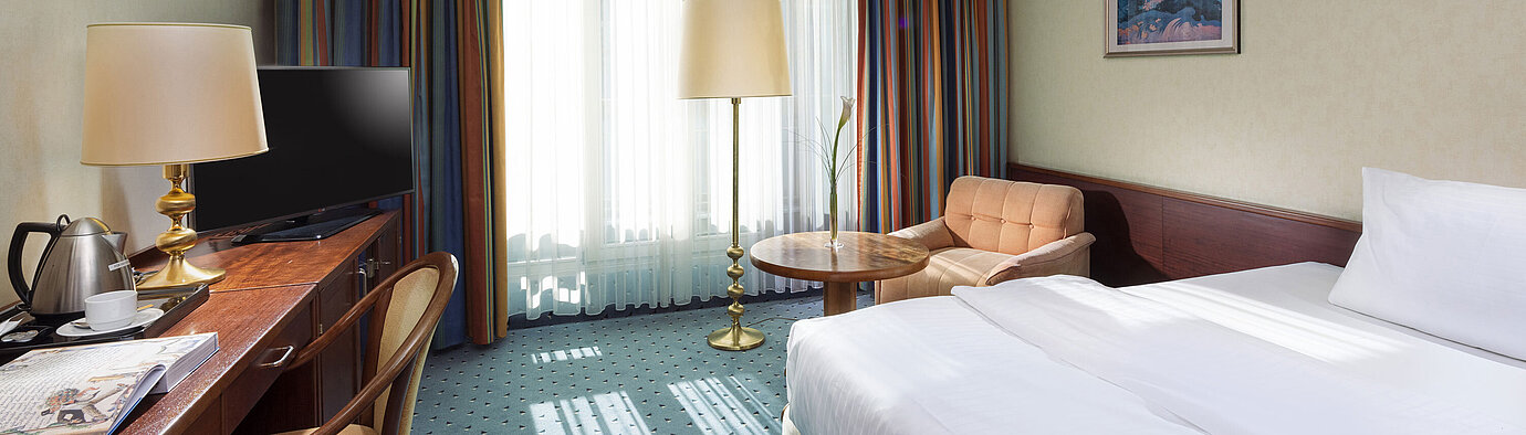 Helles Comfort Zimmer im Maritim Hotel Köln mit Einzelbett, Schreibtisch, Fernseher, Sessel und großen Fenstern mit Vorhängen