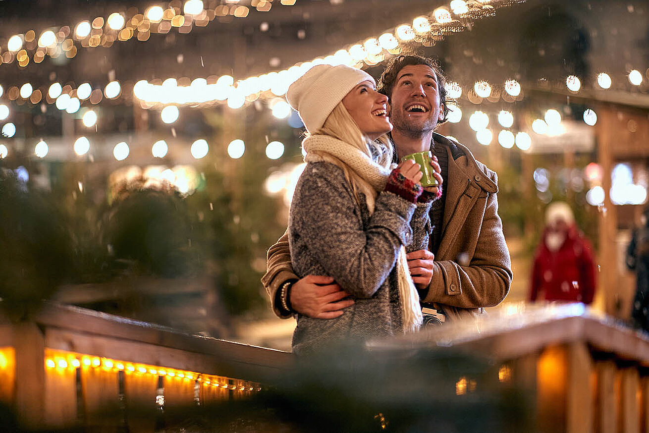 © luckybusiness - AdobeStock.com Paar genießt einen winterlichen Abend auf dem Weihnachtsmarkt, umgeben von warmen Lichtern und festlicher Stimmung.