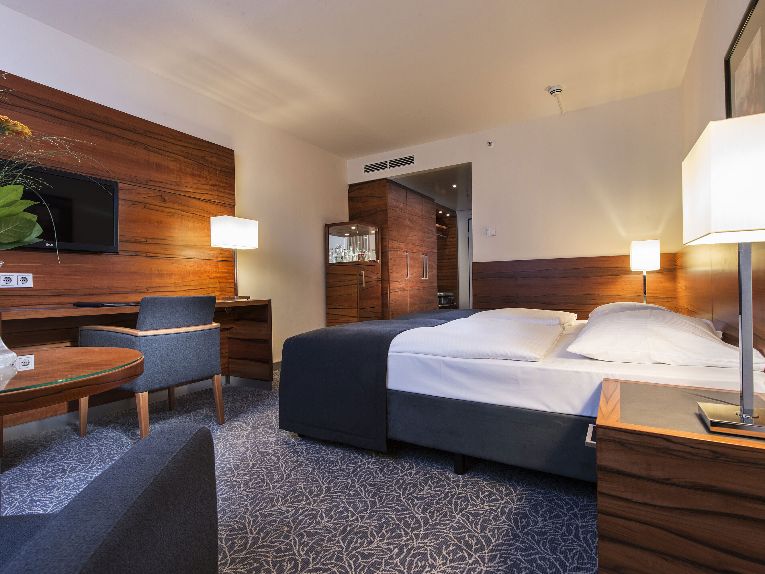 Eingerichtetes Hotelzimmer im Maritim Hotel München mit komfortablem Bett, Arbeitsbereich, TV und Holzverkleidung.