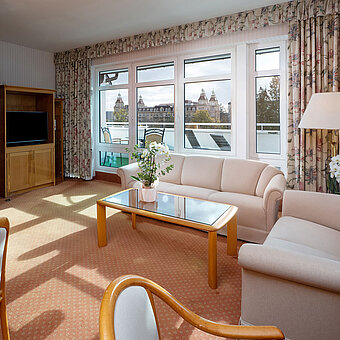 Junior Suite mit hellem Wohnbereich, Sofa, großem Fenster, Essplatz und klassischer Einrichtung im Maritim Hotel.