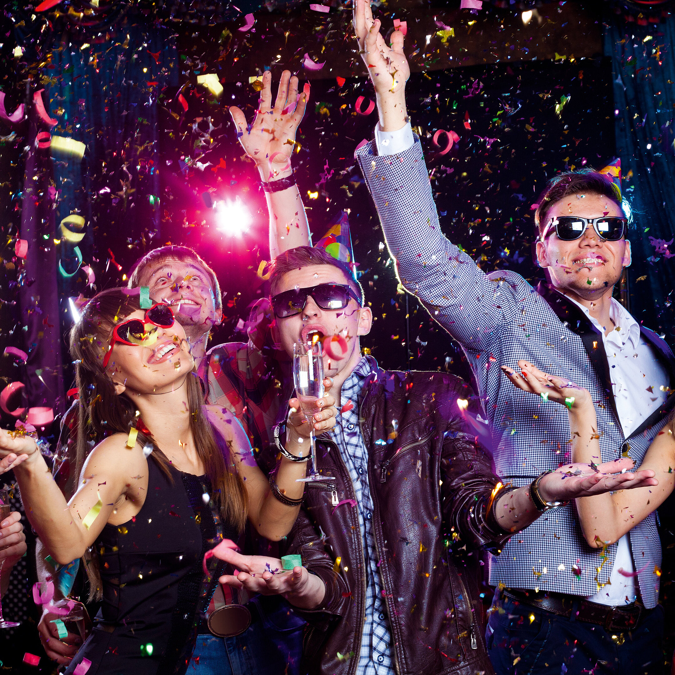 Fröhliche junge Leute feiern mit Partyhüten, Sonnenbrillen, Sektgläsern und Konfetti in bunter Clubatmosphäre