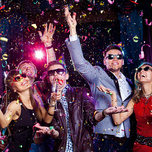 Fröhliche junge Leute feiern mit Partyhüten, Sonnenbrillen, Sektgläsern und Konfetti in bunter Clubatmosphäre