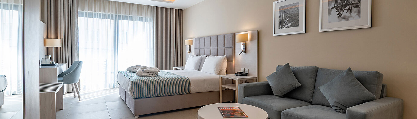 Moderne Junior Suite im Maritim Antonine Hotel & Spa Malta mit Wohnbereich, Sofa, Doppelbett und hellem Raumkonzept.