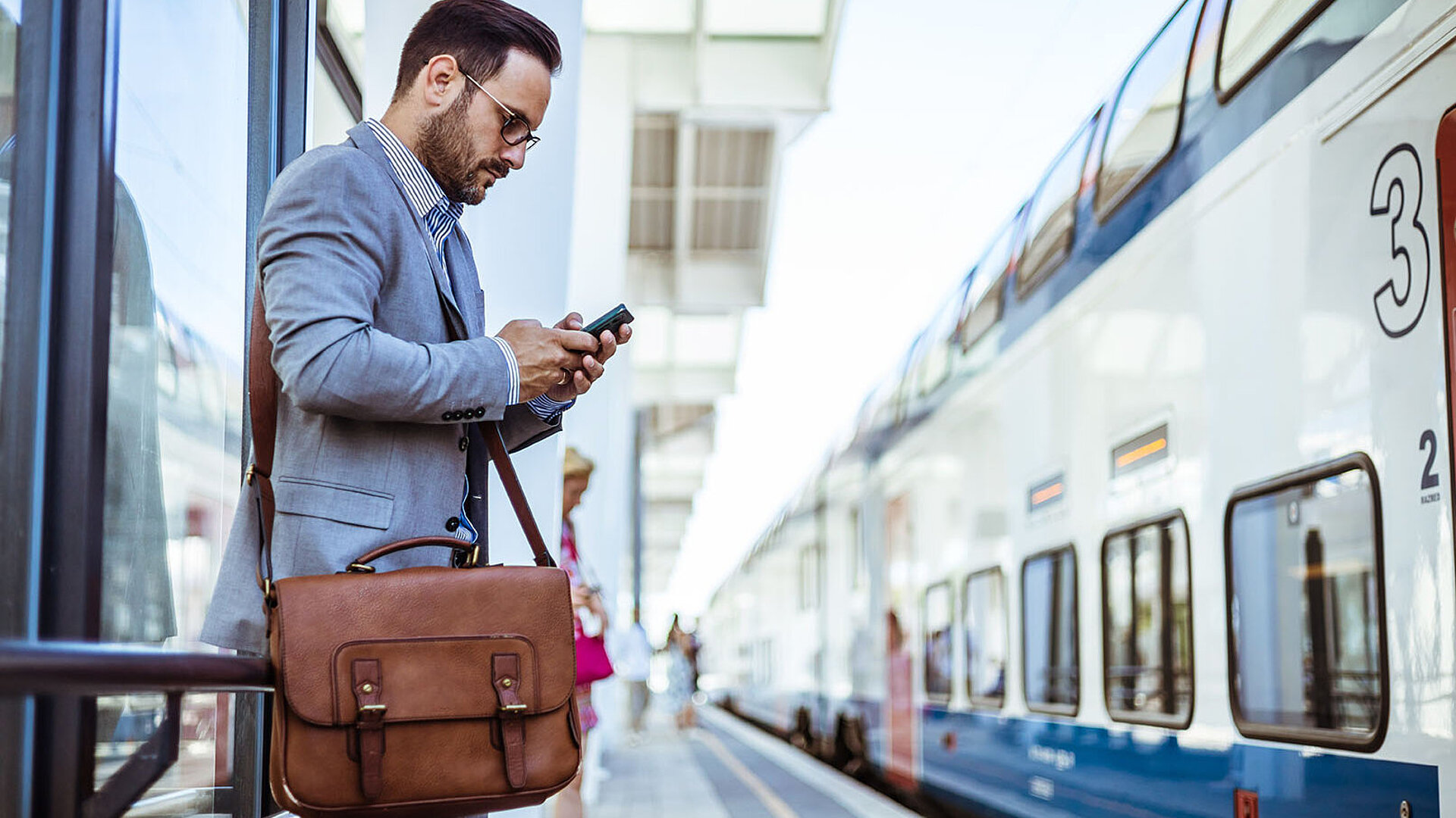 © Dragana Gordic - AdobeStock.com Geschäftsmann im Anzug mit Ledertasche wartet am Bahnsteig auf den Zug und blickt dabei auf sein Smartphone