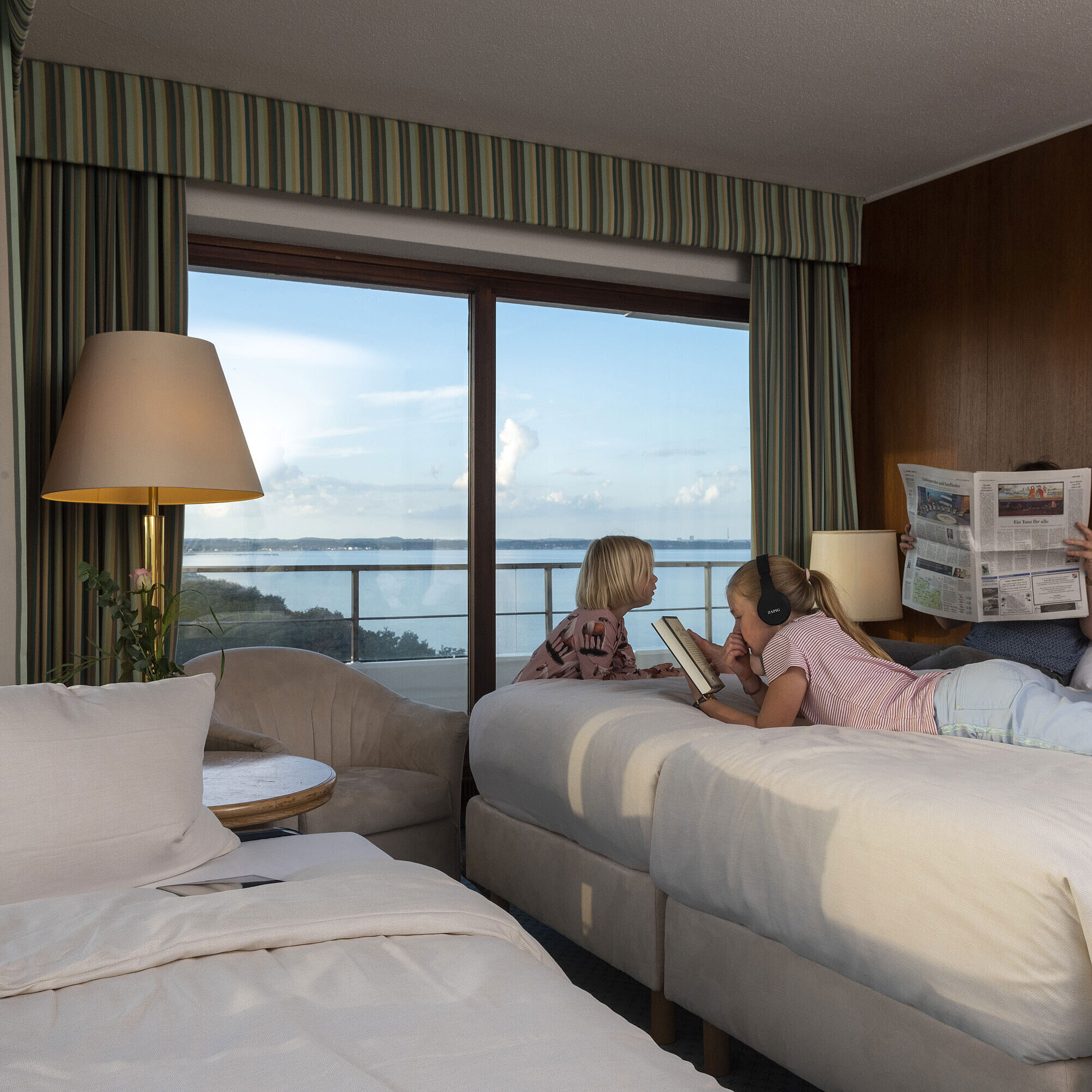 Familienzimmer im Maritim Hotel Timmendorfer Strand mit Kindern, die lesen, ein Tablet nutzen und den Meerblick genießen.