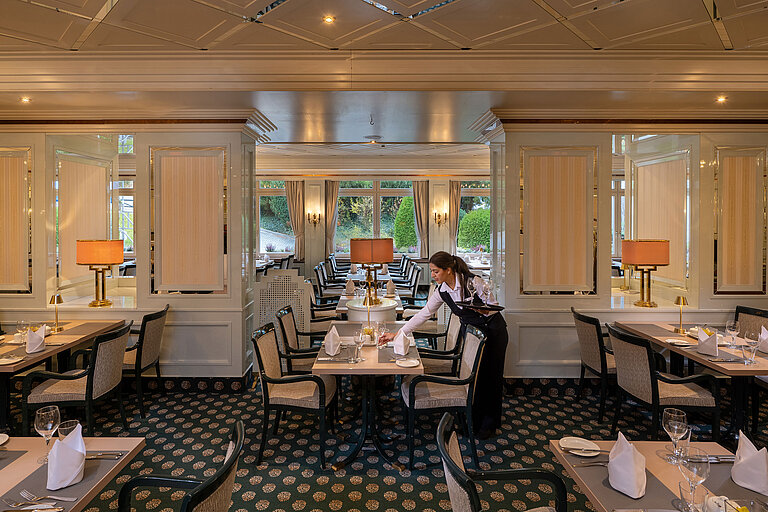 Elegantes Restaurant mit eingedeckten Tischen, große Fenster und Servicekraft beim Vorbereiten des Gastraums.