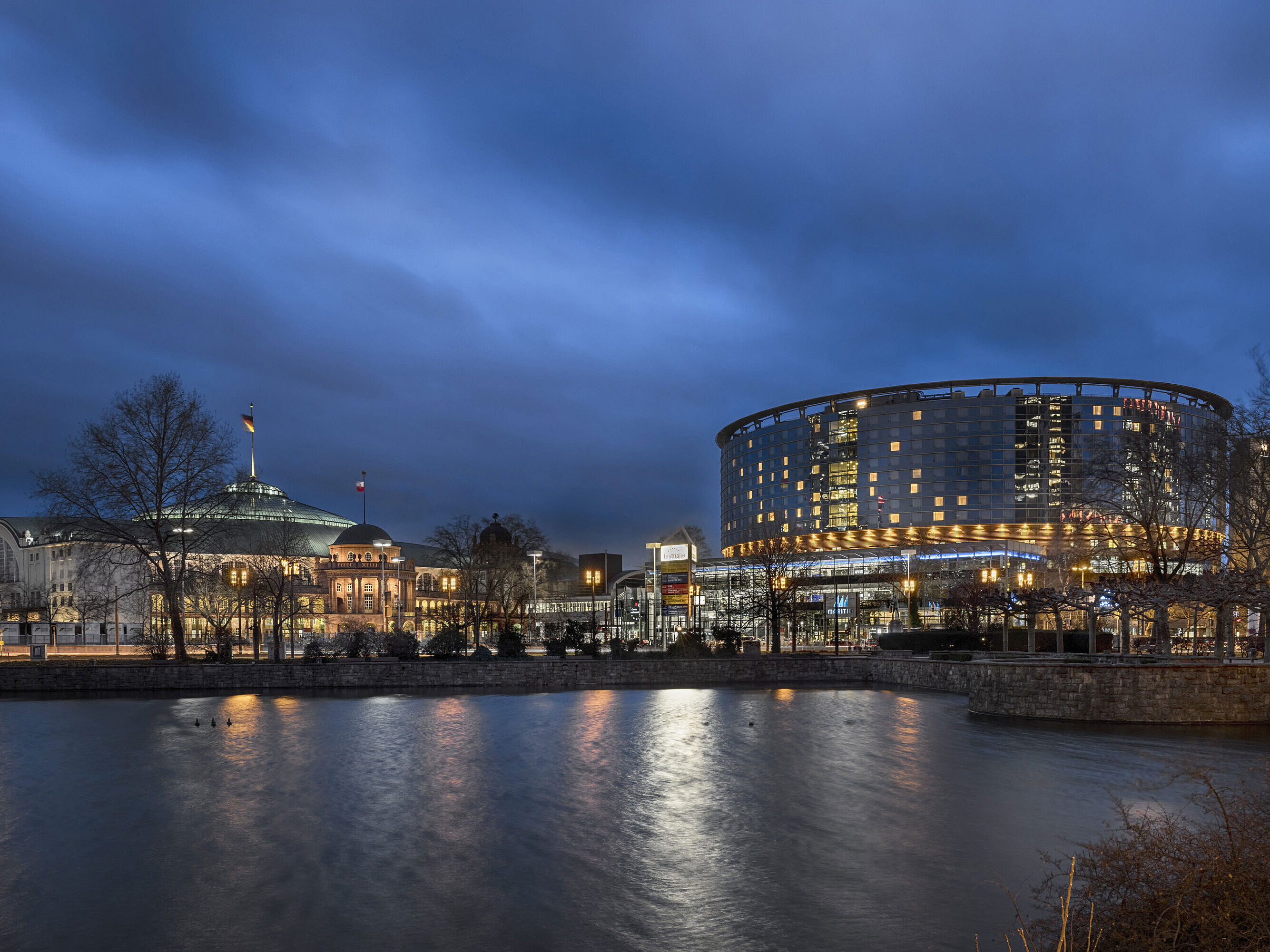 Maritim Hotel Frankfurt und Festhalle bei Nacht mit beleuchteter Glasfassade und Spiegelung im angrenzenden Teich.