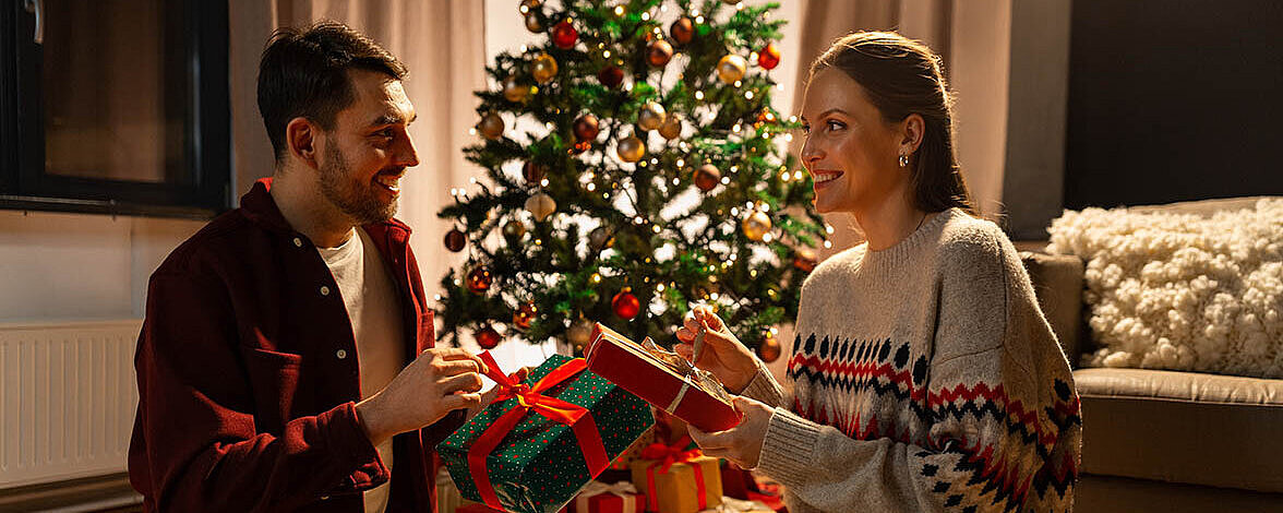 Zwei Personen sitzen vor einem geschmückten Weihnachtsbaum und tauschen liebevoll verpackte Geschenke aus.