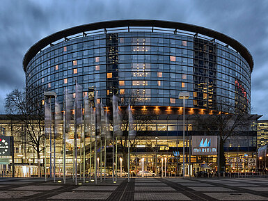 Außenansicht Frankfurt Beleuchteter Eingangsbereich des Maritim Hotel Frankfurt mit moderner Glasfassade und wehenden Flaggen bei Dämmerung.